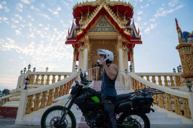 Motosikletli bir adam Tayland tapınağının önünde kask takıyor.