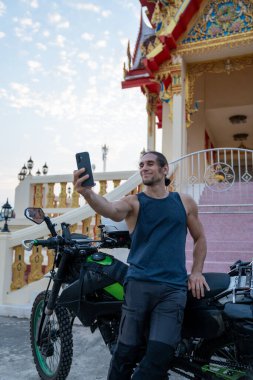 Adam gülümsüyor, akıllı telefonuyla selfie çekiyor Tayland tapınağında motosiklete yaslanıyor.