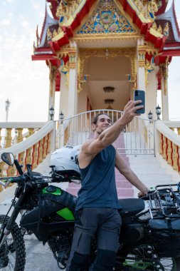 Erkek motosikletçi Tayland 'daki süslü bir Budist tapınağında akıllı telefondan seyahat videosu kaydediyor.