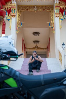 Genç adam tapınağın merdivenlerinde oturuyor, motosiklet sürüyor ve akıllı telefon kullanıyor.