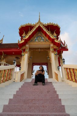 Tayland 'da mavi bir gökyüzünün altında süslü tapınak merdivenlerinde oturan, cep telefonuna bakan bir adam.