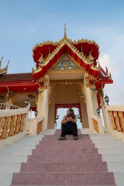 Uzun saçlı bir adam tapınak merdivenlerinde oturuyor, yukarı bakıyor, telefonu tutuyor.