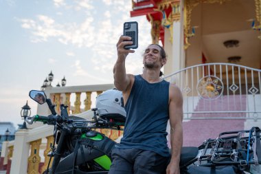 Motosikletli bir adam süslü bir Tayland tapınağının önünde selfie çekiyor.