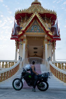Seyahat sırasında Tayland tapınağının önüne parketmiş motosikletli bir adam.