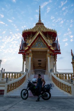 Erkek bisikletçi renkli bir Tayland Budist tapınağının önünde motosikletinin yanında duruyor.