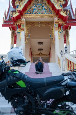 Tapınak merdivenlerinde oturan genç adam ön planda motosikletli bir telefon kullanıyor.