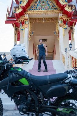 Tapınağın merdivenlerinde duran bir adam, ön tarafa park etmiş bir motosiklet, gidonun üzerinde dinlenen bir kask.