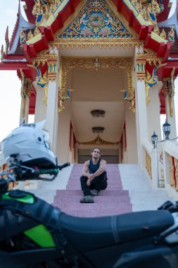 Tapınak merdivenlerinde dinlenen adam, motosiklet ve miğfer ön planda seyahat ve macerayı simgeliyor.