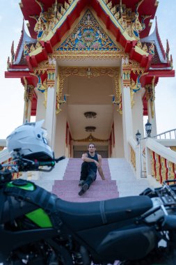 Tapınak merdivenlerinde oturan genç adam motosiklet ön planında, Tayland kültürünü keşfediyor.
