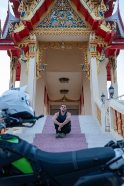 Süslü bir tapınağın önünde merdivenlerde oturan bir adam, ön planda bir motosiklet ve kask.