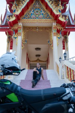 Madalyalı bir Budist tapınağının merdivenlerinde macera motosikletiyle dinlenen bir adam.