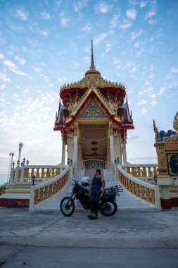 Mavi gökyüzüne karşı renkli bir Tayland Budist tapınağının önünde motosikletli bir adam duruyor.