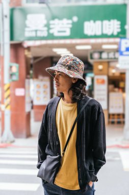 Şehir caddesinde kova şapka, hırka ve çapraz ceset torbası takan genç adam.