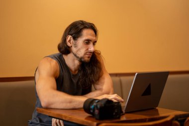 Masasında kamera olan dizüstü bilgisayar kullanan uzun saçlı bir adam bir proje üzerinde çalışıyor.