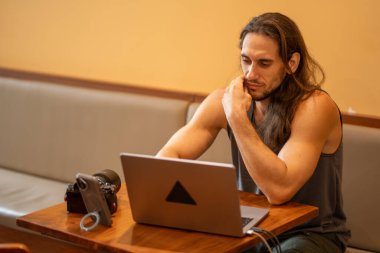 Dizüstü bilgisayara odaklanan genç adam, kamera ve telefonla içerik oluşturmayı planlıyor.