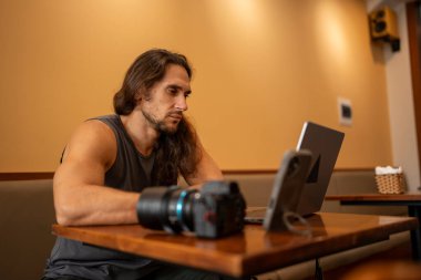 Uzun saçlı bir adam bir kafede serbest çalışıyor, kameralı ve telefonlu dizüstü bilgisayarına odaklanıyor.