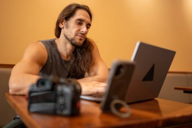 Kameralı bir kafede çalışırken dizüstü bilgisayara ve akıllı telefona odaklanan bir adam.