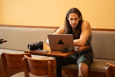 Uzun saçlı bir adam bir kafede dizüstü bilgisayarda resim düzenliyor.