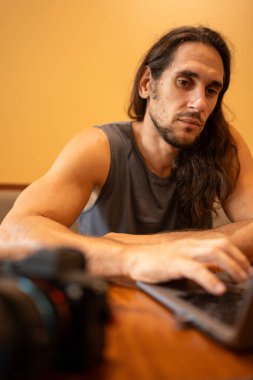 Uzun saçlı bir adam kameranın yanına dizüstü bilgisayar klavyesi yazmaya odaklanmış.