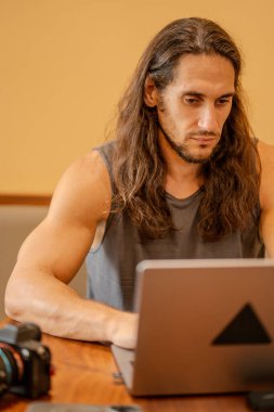Uzun saçlı bir adam dizüstü bilgisayarında daktilo kullanıyor. Yakınlarda bir yerde kamerayla çalışıyor.