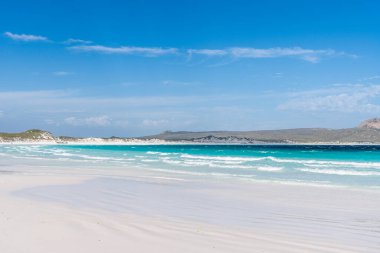 Temiz mavi gökyüzünün altında el değmemiş bir sahil, beyaz kum ve turkuaz okyanus sularının manzarası.