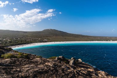Berrak turkuaz okyanus suyu ve uzak tepelerle beyaz kumsal manzarası.