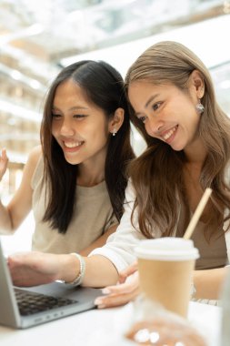 İki mutlu Asyalı kadın bir dizüstü bilgisayar ve kahve ile iş birliği yapıyor, konuşuyor ve kaynaşıyor.