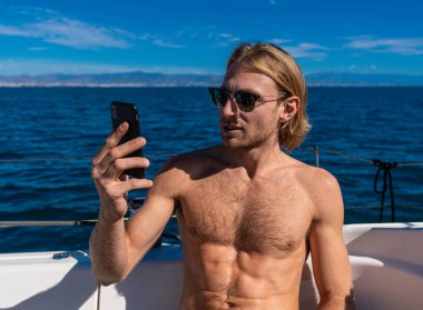 Sarışın, üstsüz, güneş gözlüklü bir adam yatta akıllı telefonuyla selfie çekiyor.