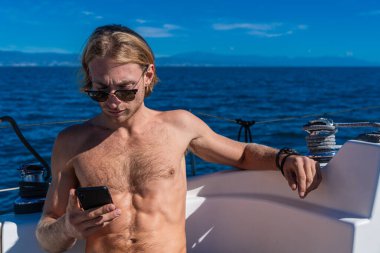 Yazın yelkenlide cep telefonuyla mesajlaşan güneş gözlüğü takan tişörtsüz bir adam.