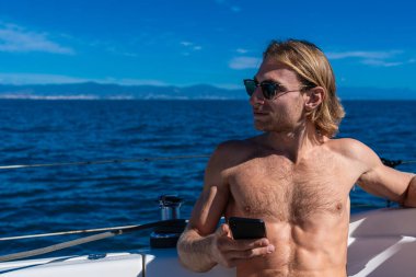 Yaz boyunca güneş gözlüğü takan ve bir teknede cep telefonu tutan genç bir adam.