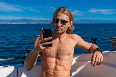 Genç adam güneş gözlüğü takıyor ve yatta akıllı telefon kullanıyor. Yaz tatilinin tadını çıkarıyor.