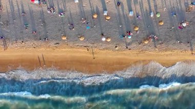 Okyanus dalgaları kıyıya vuran kumlu bir kumsalın yukarıdan aşağıya insansız hava aracı görüntüsü. Videoda sahile gidenler, şemsiyeler ve altın saat boyunca uzun gölgeler gösterilerek sakin bir yaz ortamı yaratılıyor. Kıyı şeridinin sinematik genel görünümü.