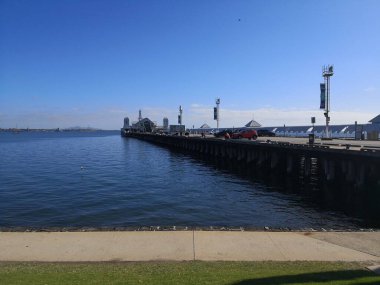 Geelong, Victoria, Avustralya - 26 Aralık 2018 - Çimenli ön plan ve uzak tepeler tarafından çerçevelenmiş binaları ve arabaları sakin suya uzanan uzun iskele.