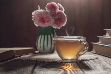 Üzerinde kitaplar ve vazoda bir buket gül bulunan bir masanın üzerinde duran, kahverengi bir fonda şeffaf bir fincan siyah çay. Çayın kalp şeklinde buharı var.