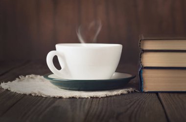 Bir fincan sıcak çay, kalp şeklinde buhar ve ahşap masa üzerinde kitap yığını, samimi okuma konsepti.