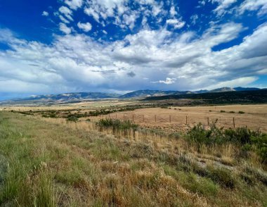 Bu fotoğraf, dinamik bulutlarla dolu yüksek bir gökyüzünün altında uçsuz bucaksız bir manzara yakalıyor. Parlak mavi gökyüzü kalın beyaz ve gri kümülüs bulutlarıyla kaplıdır..