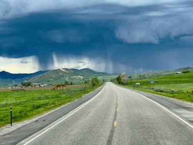 Şiddetli fırtınalar altında tepelere çıkan kırsal kesimde düz bir asfalt yol. Arka planda yerel yağış görülebilir. Fırtınanın arifesindeki manzara, tarlaların parlak yeşili kara bulutlarla çelişiyor..