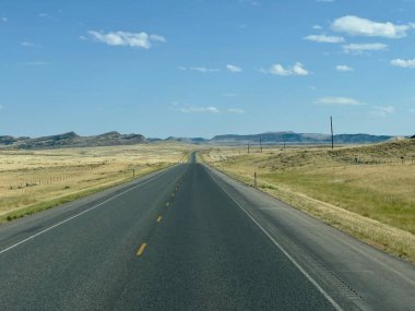Bu resim açık bir tepe boyunca ufkun çok ötesine uzanan düz bir asfalt yolu gösteriyor. Manzara, iki şeritli yolun tam ortasında sarı aralıklı işaretlerle açılıyor. Gökyüzü parlak, nazikçe mavi..