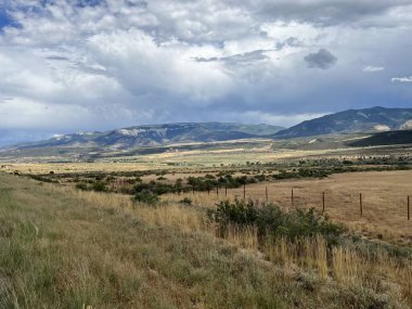 Bulutlu bir gökyüzünün altındaki kayalık bir bölgeden geçen bir yolun fotoğrafı. Sağ tarafta büyük katmanlı kumtaşı bir kaya yükselir ve gri gökyüzü yumuşak, hatta aydınlık yaratarak kanyonun sert güzelliğini vurgular..