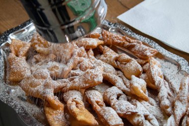 Altın kıvrımlı hamur işleri (faworki), ahşap bir tepsinin üzerinde duran parlak metal elek ile toz şekere bulanmış. Şişman Perşembe kutlamaları için çıtır çıtır, geleneksel Polonya ziyafeti, yumuşak ışık pürüzlü dokuyu ve tatlı karlı malzemeyi ön plana çıkarıyor.