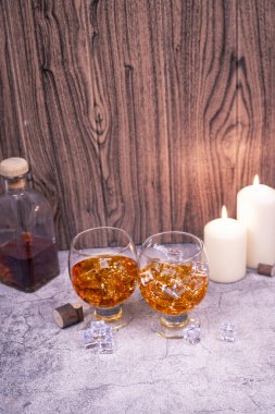 Taş bir masanın üzerinde viski ve buz olan iki kadeh, ahşap arka plan, şişe ve mumlar. Fotokopi alanı olan rahat gece içeceği konsepti.