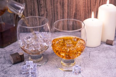 Viski dolu bir bardağın yanında buzla doldurulmuş bir bardağa dolduruluyor. Mum ışığında kırsal arka plan, bar ve bağımlılık kavramı.