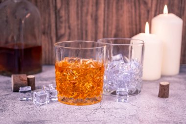 Taş bir masada iki kristal viski bardağı: biri buzlu, biri mum ve bir şişe yanında. Gece hayatı, paylaşım ya da alkolizm için rahat bir bar..