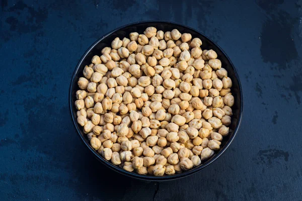 Koyu mavi arka planda siyah bir kasede pişmemiş kuru nohut manzarası. Huysuz, modern yemek..