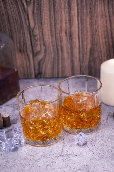 Taş bir masada buzlu iki viski bardağı, kırsal şişe ve mum ışığı arka planı. Konforlu çubuk sabit hayat, dikey kompozisyon.