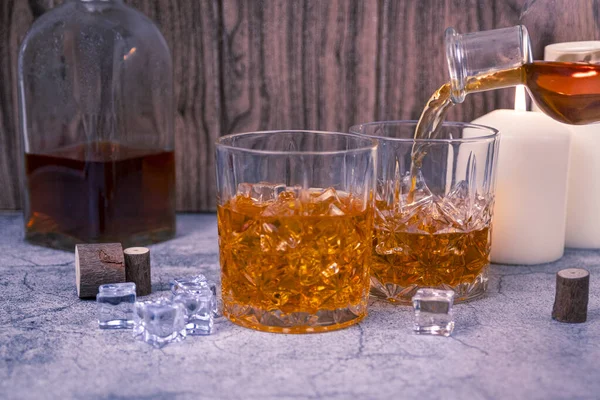 Viski ikinci bir kristal bardağa doldurulurken bir içki de şişe ve mum ışığıyla masada duruyor. Kutlama, ayartma veya alkolizm konsepti için bar servis sahnesi.