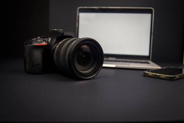 DSLR kamerası, dizüstü bilgisayarı ve akıllı telefonu olan karanlık yaratıcı çalışma alanı. İçerik oluşturma ve uzak çalışma için boş ekranlar ve kopyalama alanı.
