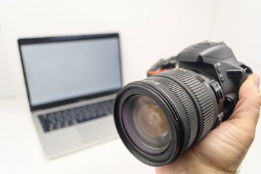 Arka planda bulanık dizüstü bilgisayarı olan bir DSLR kamera lensini tutan elin yakın çekimi, yaratıcı dijital çalışma akışı konsepti.