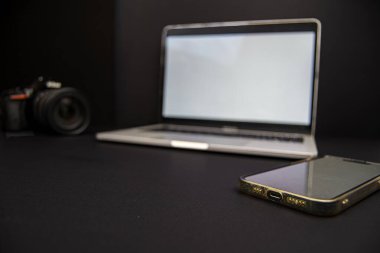 Dizüstü bilgisayarı, DSLR kamerası ve siyah arka planda akıllı telefonu olan sade bir çalışma alanı. Teknoloji, fotoğrafçılık, uzaktan çalışma ve kopyalama alanı için ideal.