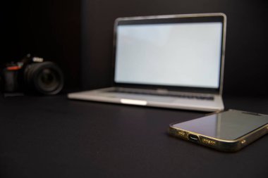 Önplanda dizüstü bilgisayar ve DSLR kamera ile yakın plan akıllı telefon koyu arkaplan, mobil iş akışı konsepti, iletişim ve yaratıcı teknoloji.
